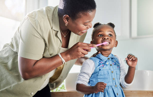 A mom helps her young child brush her teeth.